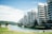 Modern apartment buildings line a waterway with several floors of balconies, surrounded by greenery and palm trees. The sky is clear with few clouds, and there is a blurred effect giving a dreamy aesthetic.