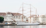 A large sailing ship with tall masts is docked beside a harbor lined with historic white buildings. The ship is adorned with colorful flags strung along its masts, and several people are seen on board. The water is calm, reflecting the ship and nearby structures.