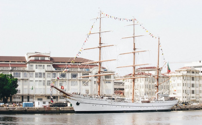 A large sailing ship with tall masts is docked beside a harbor lined with historic white buildings. The ship is adorned with colorful flags strung along its masts, and several people are seen on board. The water is calm, reflecting the ship and nearby structures.