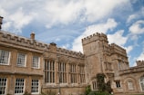 An old stone manor with tall, rectangular windows and a crenelated roofline under a bright blue sky with scattered white clouds. The architectural style is reminiscent of medieval castles and is surrounded by greenery, suggesting a peaceful rural setting.