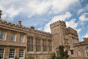 An old stone manor with tall, rectangular windows and a crenelated roofline under a bright blue sky with scattered white clouds. The architectural style is reminiscent of medieval castles and is surrounded by greenery, suggesting a peaceful rural setting.