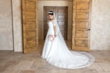 A bride stands in an elegant room, wearing a white wedding dress with a long train. She holds a bouquet of pink and white flowers and has a tiara on her head. The background features rustic wooden doors and soft, neutral walls, creating a warm and inviting atmosphere.