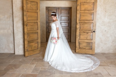 A bride stands in an elegant room, wearing a white wedding dress with a long train. She holds a bouquet of pink and white flowers and has a tiara on her head. The background features rustic wooden doors and soft, neutral walls, creating a warm and inviting atmosphere.