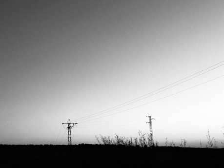 A black and white landscape features two electricity pylons connected by power lines stretching across an open area with low vegetation in the foreground. The gradient sky transitions from a lighter to darker shade of gray towards the top.