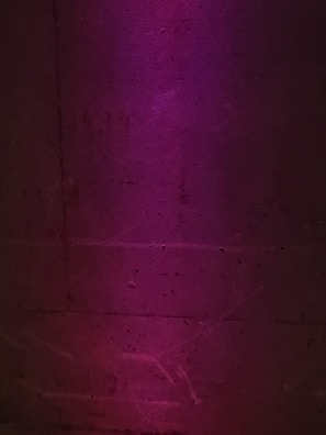 A roughly textured concrete wall illuminated by a purple light, creating subtle patterns and shadows.