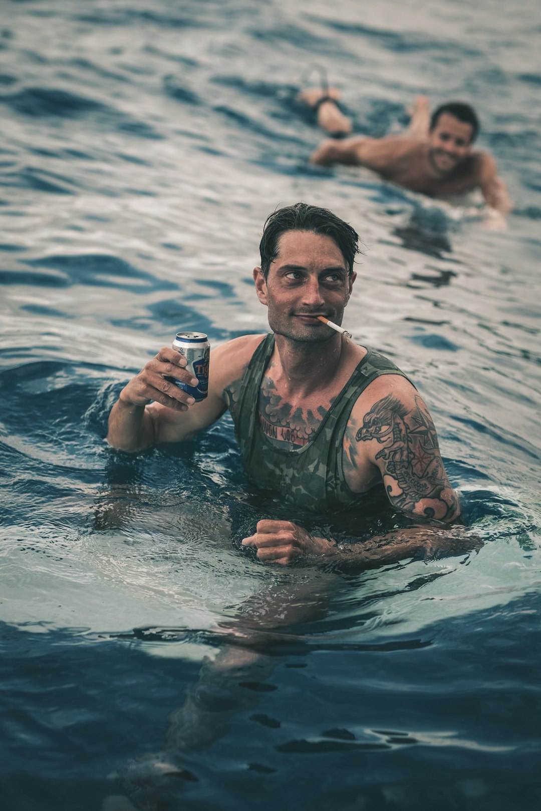 Two soldiers swimming, one with a cigarette in his mouth and a can of beer in his hand.