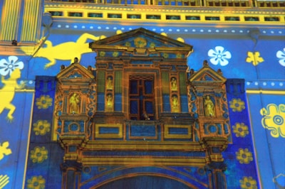 A colorful projection on a classic architectural facade with intricate patterns in blue, gold, and yellow, showcasing decorative elements and floral motifs. The artwork includes images of horses and flowers, adding a vibrant and artistic touch.