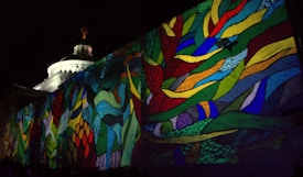 A vibrant projection of multicolored abstract patterns covers a large outdoor wall, with a classical dome structure partially visible in the background. The design consists of intertwining shapes and lines in vivid colors such as red, green, blue, and yellow against the nighttime sky.