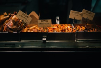 A display of various roasted foods including rotisserie chicken, roasted potatoes, roast cauliflower with harissa, and roasted carrots with yogurt and tahini. Each dish is separated into metal trays with signs indicating their names and descriptions.