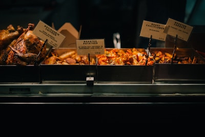 A display of various roasted foods including rotisserie chicken, roasted potatoes, roast cauliflower with harissa, and roasted carrots with yogurt and tahini. Each dish is separated into metal trays with signs indicating their names and descriptions.