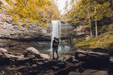 A couple stands embracing near a waterfall in a forest setting. The waterfall cascades over a rocky cliff, with abundant greenery and trees surrounding the area. Large rocks are scattered across the foreground, adding to the natural ambiance.
