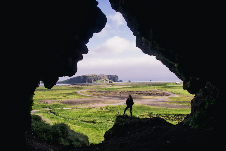 The image depicts a person standing on a cliff, overlooking a vast landscape below. The individual is positioned near the center of the frame and appears to be gazing out at the horizon. The scene is set against a backdrop of lush green fields that stretch out into the distance, with a body of water visible in the background. The sky above is filled with clouds, adding depth and texture to the image.