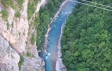 Aerial view of a winding river cutting through a lush green forest with steep rocky cliffs on one side. A small raft with people is visible on the river, suggesting white-water rafting activity. The water appears clear and turquoise, contrasting with the dense foliage and rocky terrain.