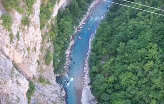 Aerial view of a winding river cutting through a lush green forest with steep rocky cliffs on one side. A small raft with people is visible on the river, suggesting white-water rafting activity. The water appears clear and turquoise, contrasting with the dense foliage and rocky terrain.