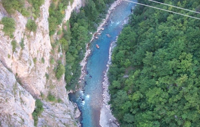 Aerial view of a winding river cutting through a lush green forest with steep rocky cliffs on one side. A small raft with people is visible on the river, suggesting white-water rafting activity. The water appears clear and turquoise, contrasting with the dense foliage and rocky terrain.