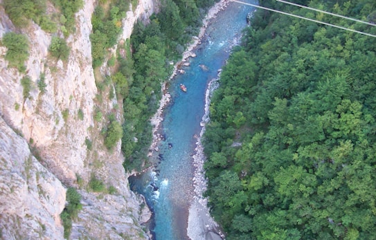 Aerial view of a winding river cutting through a lush green forest with steep rocky cliffs on one side. A small raft with people is visible on the river, suggesting white-water rafting activity. The water appears clear and turquoise, contrasting with the dense foliage and rocky terrain.