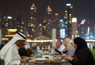 An elegant group dining alfresco under twinkling lights with a backdrop of a stunning city skyline.