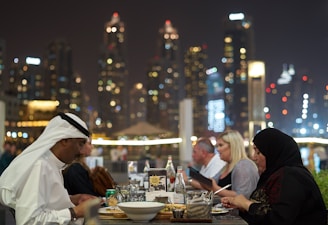 A group of people are dining at an outdoor restaurant with a view of illuminated skyscrapers in the background. The setting suggests an upscale environment, possibly in an urban city. The diners are enjoying a meal, with various dishes and beverages spread across the table.