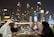 A group of people are dining at an outdoor restaurant with a view of illuminated skyscrapers in the background. The setting suggests an upscale environment, possibly in an urban city. The diners are enjoying a meal, with various dishes and beverages spread across the table.