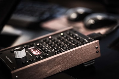 An electronic device with a wooden frame, featuring a control panel with buttons and a white knob. The device displays a red digital reading and is positioned on a dark, blurred background.