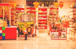 A colorful shop interior filled with a variety of items, including numerous shelves filled with decorative products. At the center is a vibrant auto-rickshaw adorned with Bollywood-themed artwork. The shop is adorned with hanging lamps and there's an array of products on display including trinkets, textiles, and ornaments.