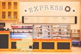 A cozy café interior featuring a display counter filled with various pastries and bottled drinks. Above the counter, a large illuminated sign reads 'EXPRESSO' adorned with stars. Behind the counter, there are shelves with products like small jars and bottles. The menu on the wall lists various beverages. In the foreground, there's a small table with a mint-green chair.