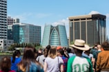 A modern cityscape featuring distinctive glass buildings in the background, surrounded by a diverse group of people in the foreground. The central building has a unique architectural design with a curved glass facade.
