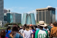 A modern cityscape featuring distinctive glass buildings in the background, surrounded by a diverse group of people in the foreground. The central building has a unique architectural design with a curved glass facade.