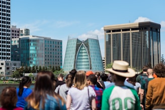 A modern cityscape featuring distinctive glass buildings in the background, surrounded by a diverse group of people in the foreground. The central building has a unique architectural design with a curved glass facade.