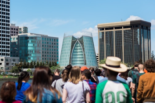 A modern cityscape featuring distinctive glass buildings in the background, surrounded by a diverse group of people in the foreground. The central building has a unique architectural design with a curved glass facade.