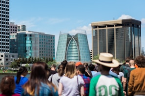 A modern cityscape featuring distinctive glass buildings in the background, surrounded by a diverse group of people in the foreground. The central building has a unique architectural design with a curved glass facade.