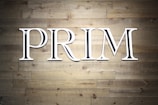 Large white capital letters spelling the word 'PRIM' are mounted against a textured wooden wall. The wall is made up of horizontal wooden planks with a natural grain pattern.