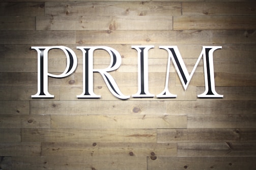 Large white capital letters spelling the word 'PRIM' are mounted against a textured wooden wall. The wall is made up of horizontal wooden planks with a natural grain pattern.
