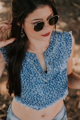 A person with long dark hair wearing sunglasses and a blue patterned sleeveless top with lace edges. The top has a decorative lace-up design near the neckline and is paired with light-colored denim shorts. The person is accessorized with teardrop-shaped earrings.