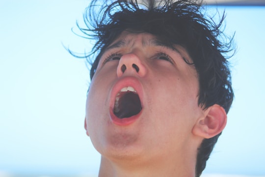 A young person with an open mouth and eyes looking upwards, with wind-blown hair and a bright background.