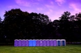 A row of portable toilets stands on a grassy field with a backdrop of large trees. The sky above is filled with vibrant colors from a sunset or sunrise, featuring shades of purple and pink.