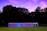 A row of portable toilets stands on a grassy field with a backdrop of large trees. The sky above is filled with vibrant colors from a sunset or sunrise, featuring shades of purple and pink.