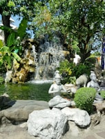 A serene garden scene features a small waterfall cascading over rocks into a pond. Several statues of meditative figures are positioned around the water, surrounded by lush green foliage. The setting is peaceful and invites contemplation.
