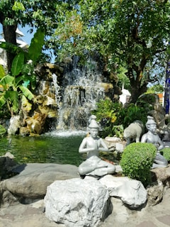 A serene garden scene features a small waterfall cascading over rocks into a pond. Several statues of meditative figures are positioned around the water, surrounded by lush green foliage. The setting is peaceful and invites contemplation.
