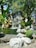 A serene garden scene features a small waterfall cascading over rocks into a pond. Several statues of meditative figures are positioned around the water, surrounded by lush green foliage. The setting is peaceful and invites contemplation.