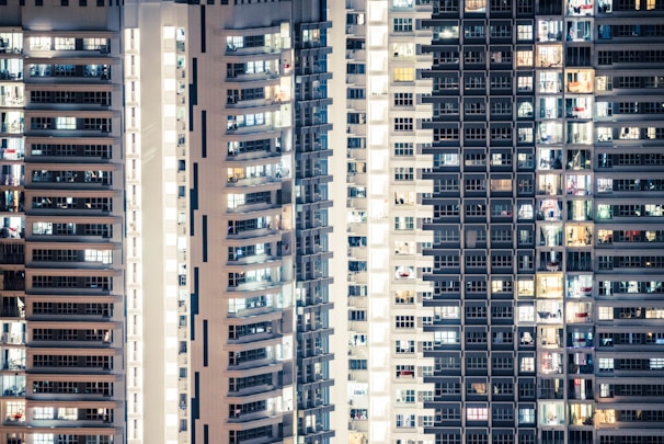 High-rise apartment buildings with numerous windows, some illuminated and others dark, creating a pattern of light and shadow. The structures appear to be densely packed, showcasing urban residential living.