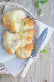 A rustic loaf of braided bread is positioned on a wicker tray with a piece of white paper towel underneath. The bread displays a golden-brown crust with visible sugar granules and is garnished with small white flowers. A knife with a white handle lies next to the bread, and small green leaves and flowers are scattered around. The background features a wooden surface that enhances the natural and cozy atmosphere.