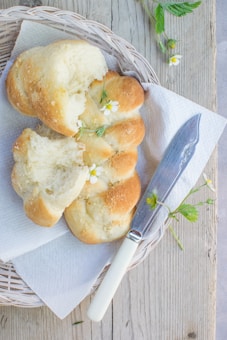 A rustic loaf of braided bread is positioned on a wicker tray with a piece of white paper towel underneath. The bread displays a golden-brown crust with visible sugar granules and is garnished with small white flowers. A knife with a white handle lies next to the bread, and small green leaves and flowers are scattered around. The background features a wooden surface that enhances the natural and cozy atmosphere.