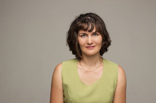 A woman with short, curly brown hair wearing a sleeveless green dress against a plain gray background. She is looking directly at the camera with a faint smile.
