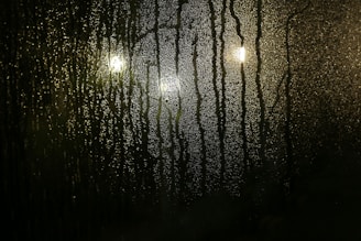 Condensation covers a glass surface, with water droplets streaming downwards. Three blurred lights are visible behind the glass, casting a soft glow and adding to the misty appearance.