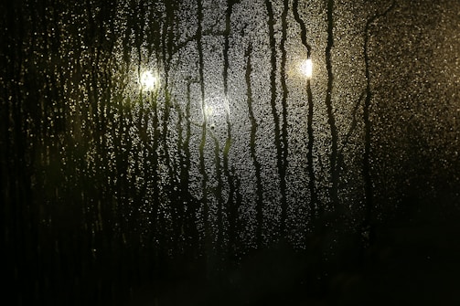 Condensation covers a glass surface, with water droplets streaming downwards. Three blurred lights are visible behind the glass, casting a soft glow and adding to the misty appearance.