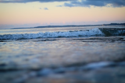 The image captures gentle ocean waves rolling towards the shore under a pastel-colored sky. The horizon features distant landforms, suggesting a coastal scene. Soft clouds are scattered in the sky, adding to the tranquil atmosphere.