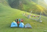 A group of people stands on a grassy hillside near three camping tents. Colorful prayer flags are strung between poles, adding vibrancy to the scene. The background features rolling hills under a clear sky, creating a serene and natural setting.