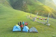 A group of people stands on a grassy hillside near three camping tents. Colorful prayer flags are strung between poles, adding vibrancy to the scene. The background features rolling hills under a clear sky, creating a serene and natural setting.