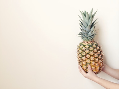 A large pineapple is being held by two hands against a plain, light-colored background. The pineapple is ripe, with a mix of green and yellow hues on its skin and a crown of spiky green leaves on top.
